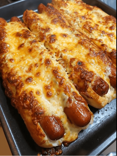 A vertical, high-angle close-up of three hot dogs in buns, tightly packed in a dark baking pan. The hot dogs are buried under a thick, bubbly layer of melted golden-brown cheese flecked with green herbs. The tips of the browned, juicy franks peek out from the ends of the toasted buns. The lighting is bright and warm, emphasizing the crispy texture of the cheese.