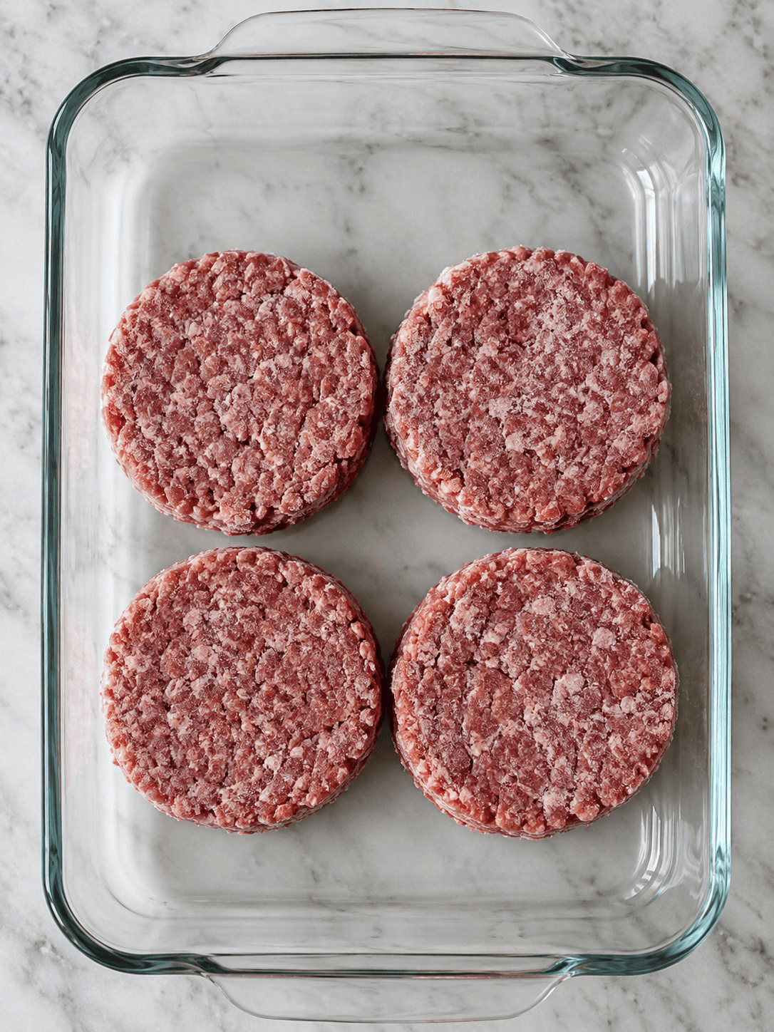 Four frozen raw beef patties in a clear glass baking dish