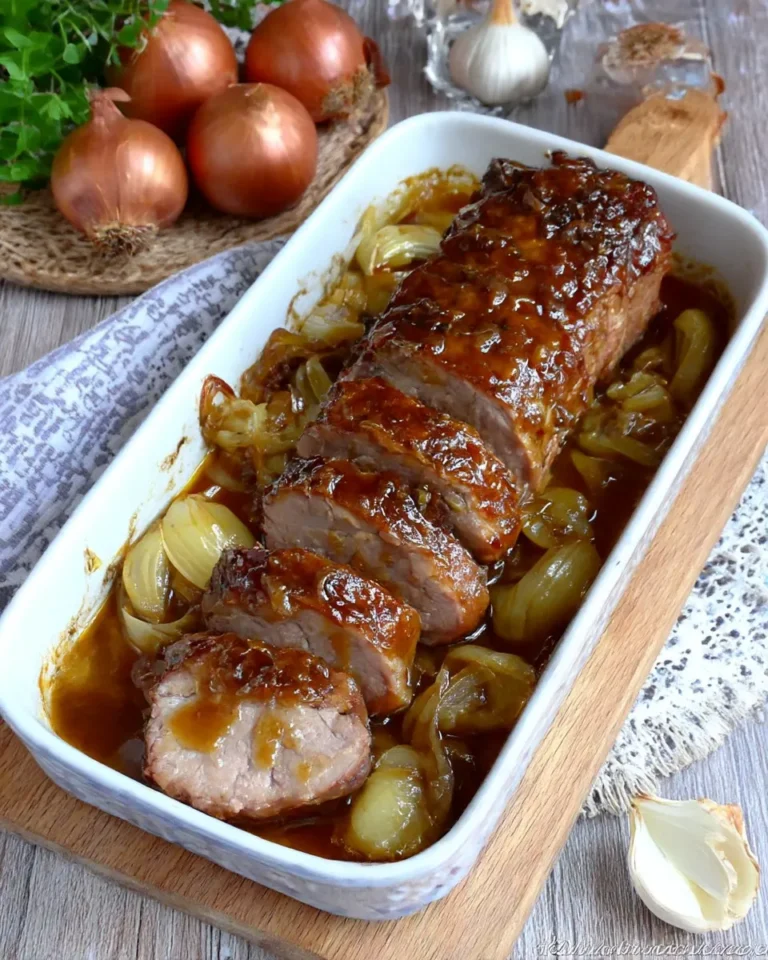 A top-down, vertical shot of a sliced pork loin roast in a white rectangular ceramic baking dish. The pork is covered in a thick, glistening honey-garlic glaze and surrounded by soft, golden caramelized onion wedges. The meat is tender and juicy inside, and the background features whole raw onions and a head of garlic on a rustic wooden surface.