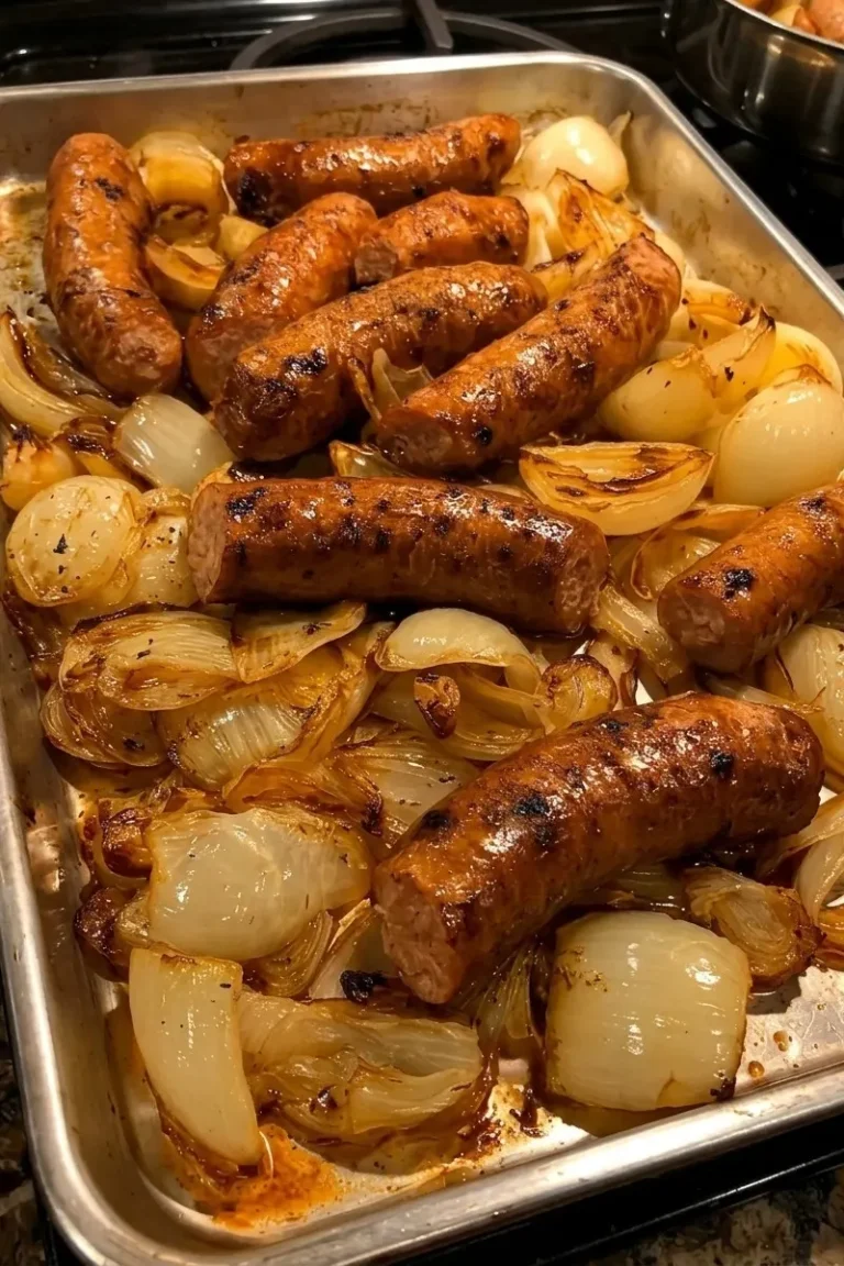 A vertical, top-down shot of a silver rimmed baking sheet filled with roasted smoked sausages and caramelized onions. The sausages are large, whole links with a deep, bronzed, and slightly charred skin. They are nestled among thick, translucent yellow onion wedges that have turned golden-brown and jammy from roasting. The pan shows savory brown juices and bits of caramelization, suggesting a rich, savory flavor.