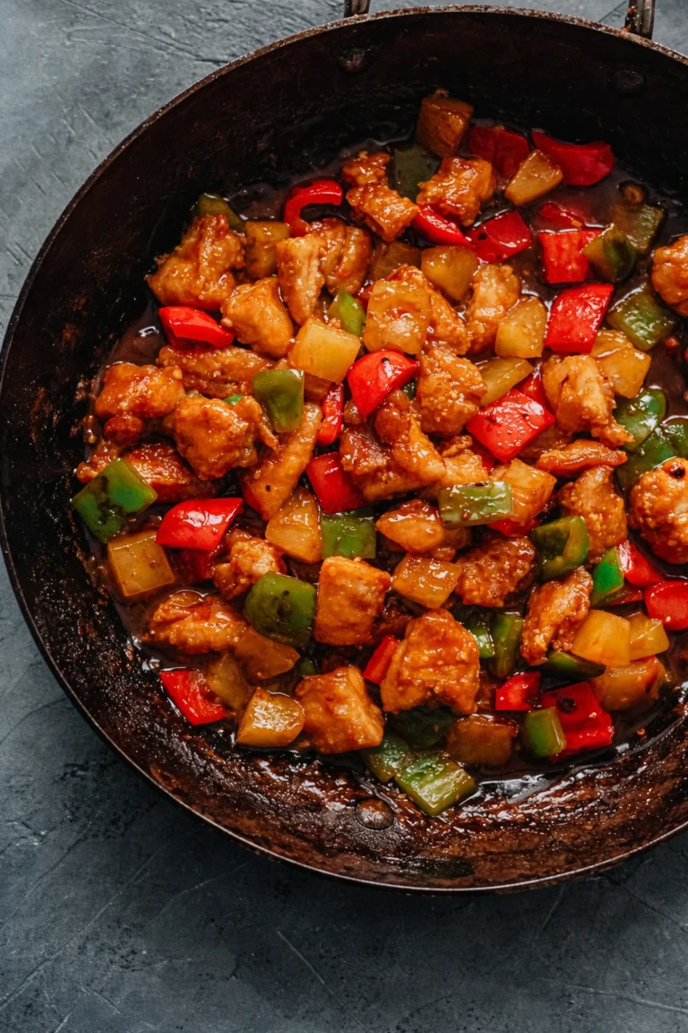 A vertical, overhead shot of a dark, seasoned wok filled with a glistening pineapple chicken stir-fry. The dish features golden-brown chicken pieces, vibrant red and green bell pepper squares, and yellow pineapple chunks, all coated in a rich, dark glossy sauce. A large dark wooden spoon is partially submerged in the wok, ready to serve.