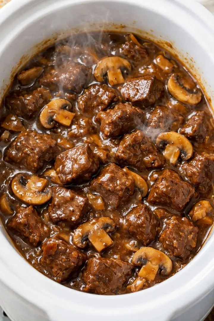 Tender slow cooker beef tips and mushrooms in rich brown gravy served hot in a white crockpot